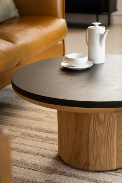 Round coffee table with dark coffee ground surface and natural wood base, styled with glass teapot and cups on beige sofa background
